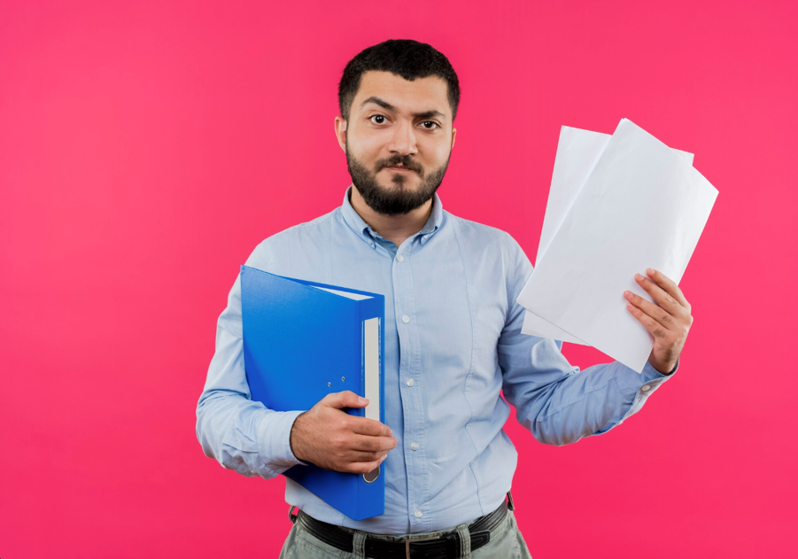 Young bearded man in blue shirt holding folder and blank pages displeased